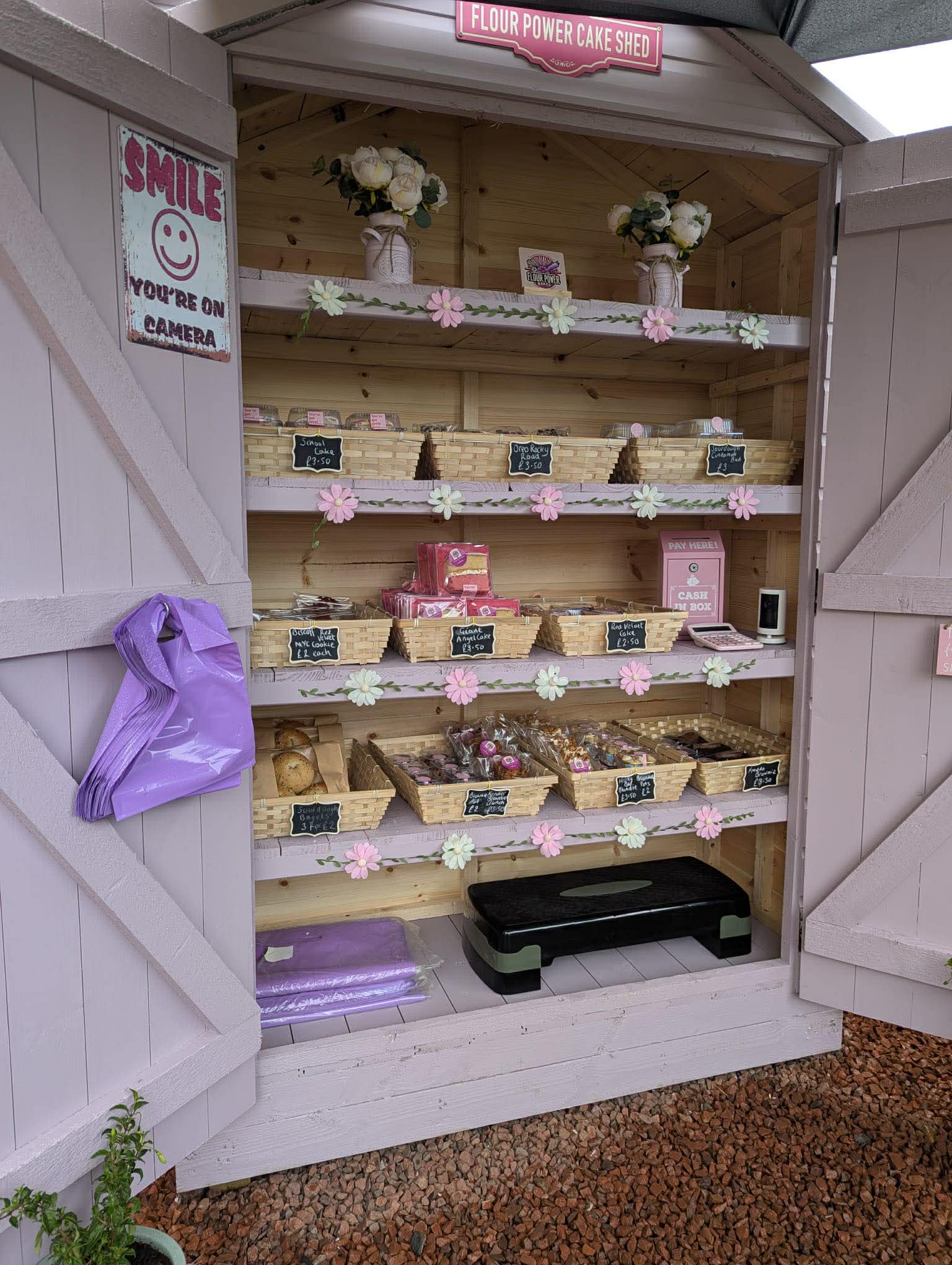 The Honesty Cake Shed with its doors open, shelves stocked with baskets of treats